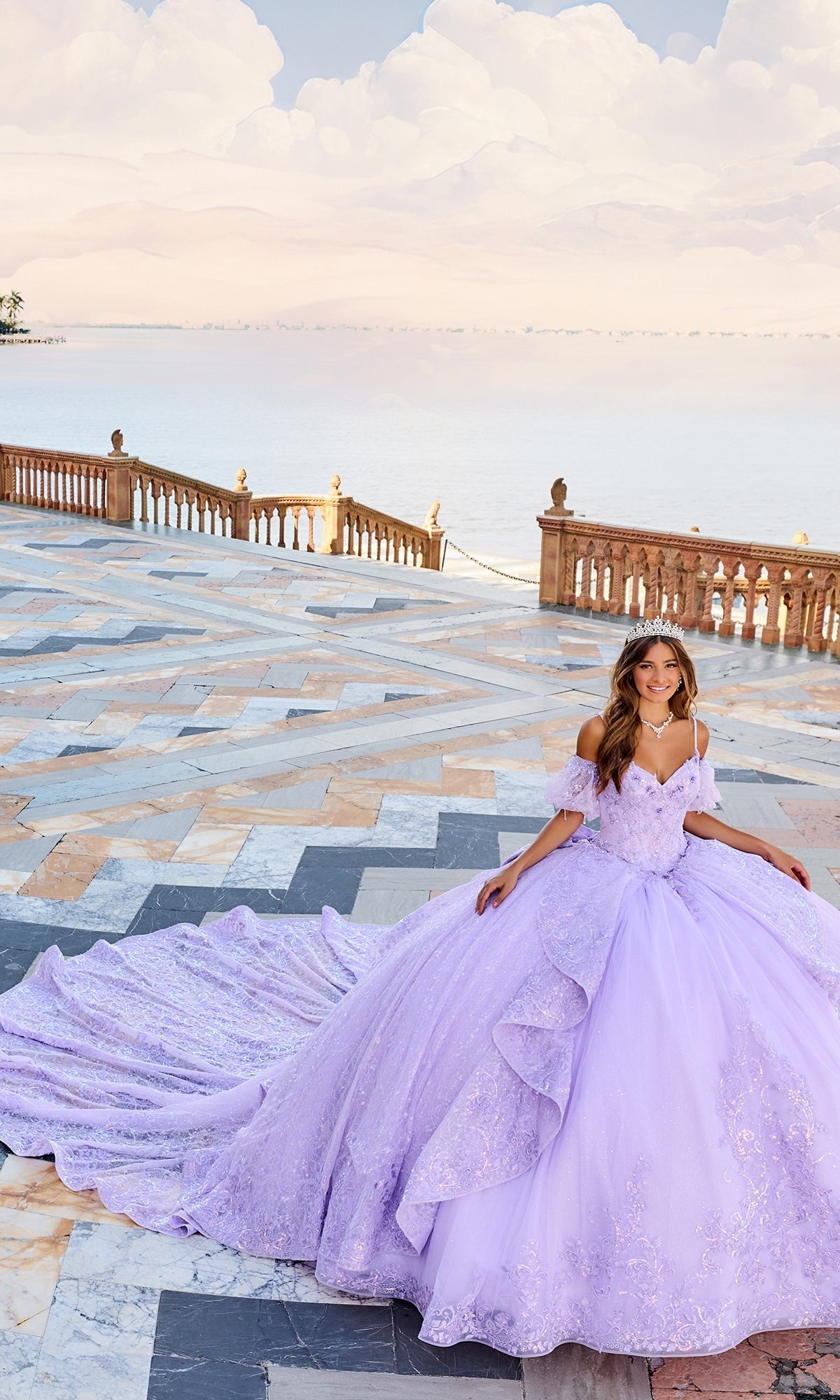 A woman wearing the Lace Quinceanera Dress: Princesa PR30161, a lavender lace gown with a detachable train, stands on a marble terrace by the water, smiling and wearing a tiara.
